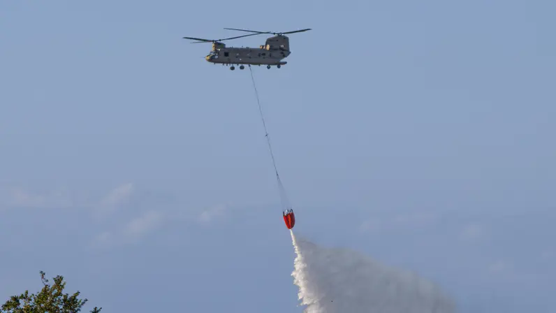 Een helikopter hangt een rode emmer uit, waaruit water wordt gesprayd op een bosbrand onder een heldere hemel.