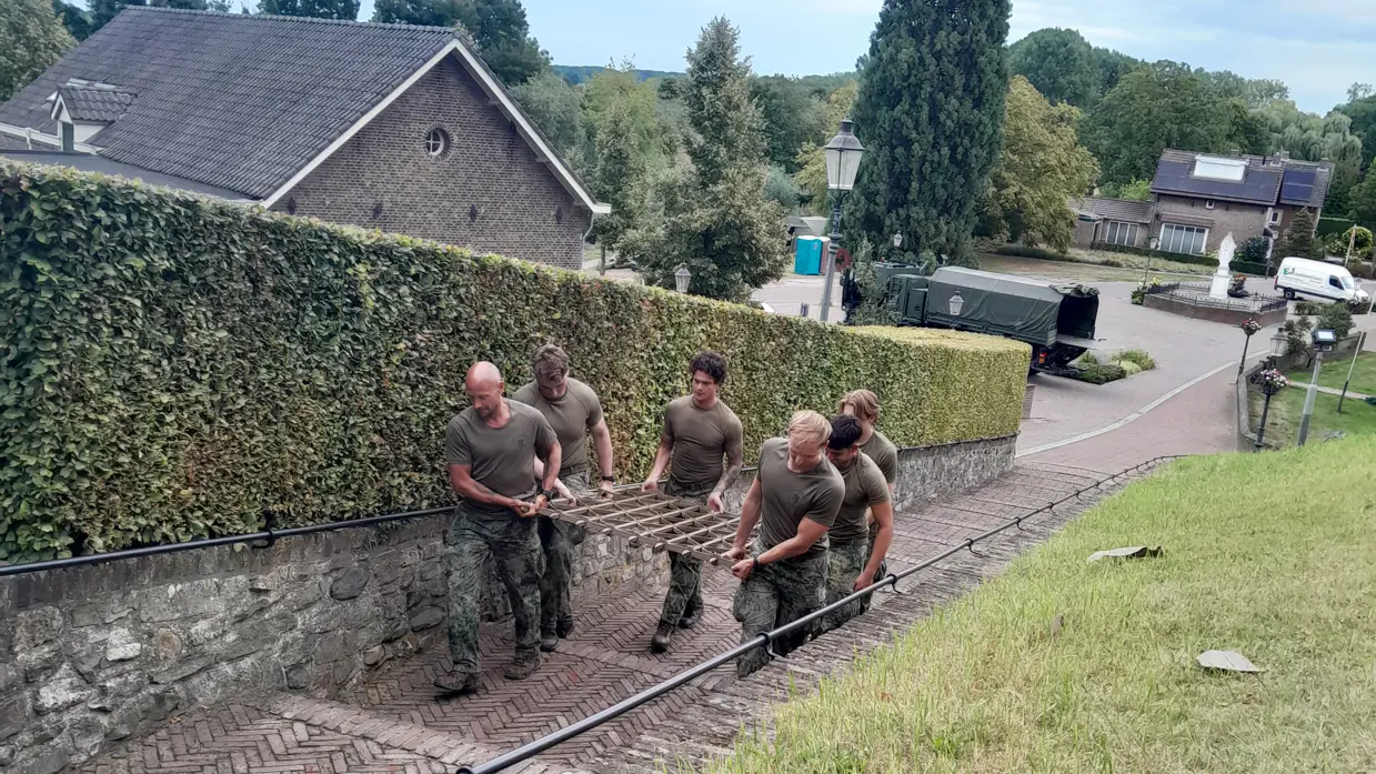 Vier militairen beklimmen een steile, begroeide helling langs een smal pad met een lage stenen muur en bomen.