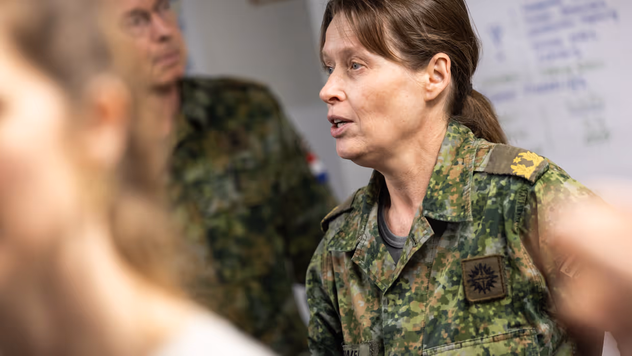 Links: Drie militairen in uniform staan in gesprek. Een man wijst uitleggend naar voren, een vrouw luistert aandachtig, en een andere man staat met gekruiste armen. Rechts: Een vrouw in militair uniform met naam praat serieus tijdens een bespreking. Op de achtergrond staan collega's in uniform bij een whiteboard. 