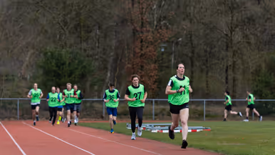 Een groep hardlopers in gele hesjes met nummers rent op een atletiekbaan. De voorste loper lijkt gefocust, met bomen en een hek op de achtergrond.