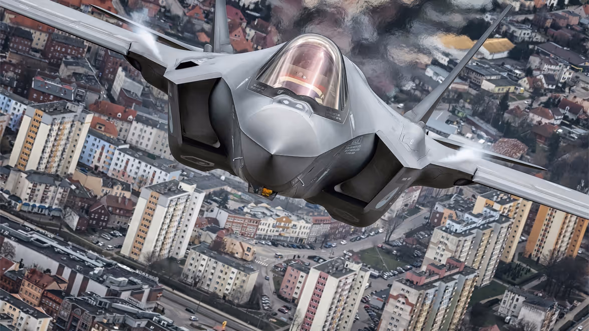 Een straaljager vliegt omhoog boven een stad met dicht opeen gebouwde flats, huizen en wegen.