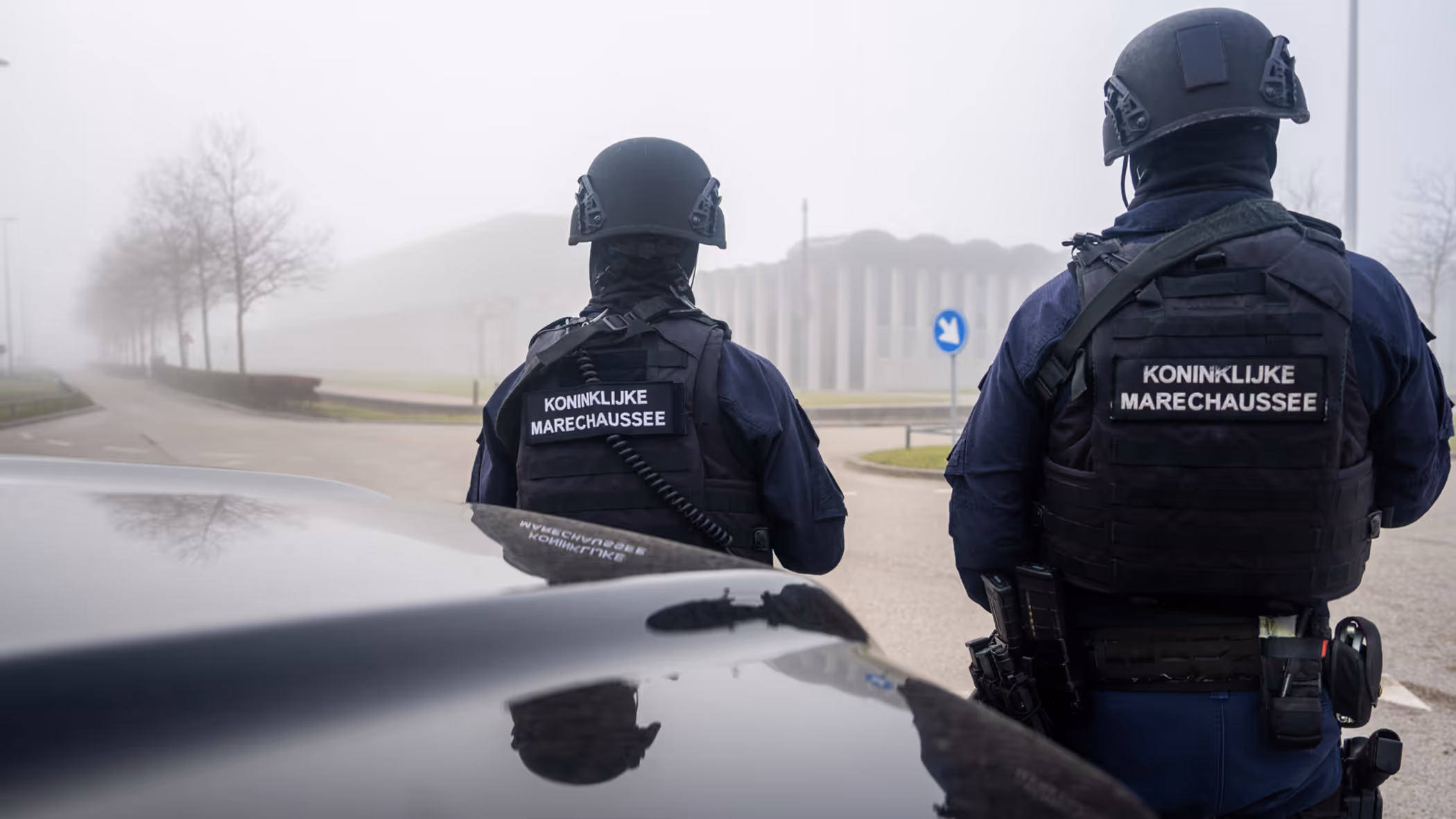 Twee zwaarbewapende marechaussees staan met hun rug naar je toe bij een auto op een mistige, rustige weg.