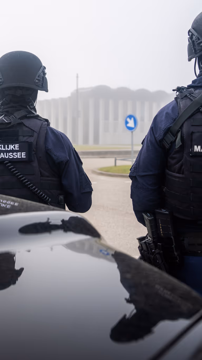 Twee zwaarbewapende marechaussees staan met hun rug naar je toe bij een auto op een mistige, rustige weg.