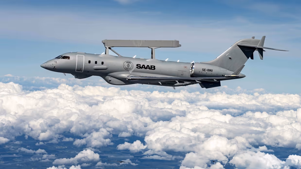 Een grijs militair vliegtuig van Saab vliegt hoog boven de wolken met een grote radarbobbel op de rug en registratie SE-RMV op de staart. 