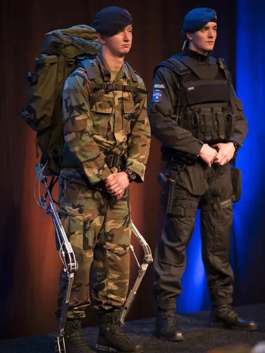 Links: Twee mannen in uniform: één met camouflage, rugzak en exoskelet, de andere in zwart met kogelwerend vest.    Rechts: Een man in grijs jack praat met een soldaat in camouflage en rugzak in een groen bos. 