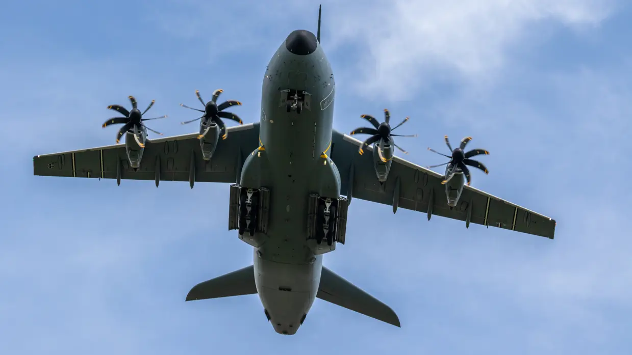  Een grijs militair transportvliegtuig met vier propellers vliegt laag tegen een blauwe hemel met lichte wolken. 