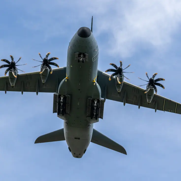  Een grijs militair transportvliegtuig met vier propellers vliegt laag tegen een blauwe hemel met lichte wolken. 