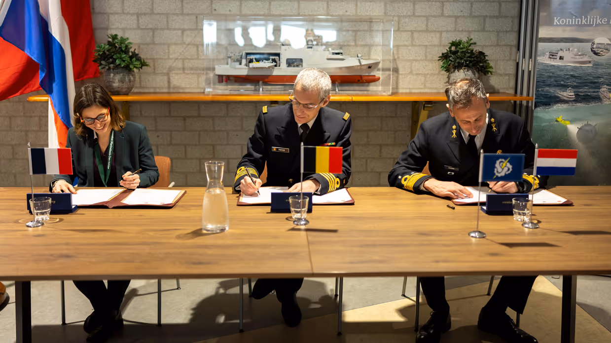 Een sprekende vice-admiraal Jan Willem Hartman, daarnaast zit hij met een burger en een andere militair achter en tafel en zetten zij gelijktijdig een handtekening onder een document.