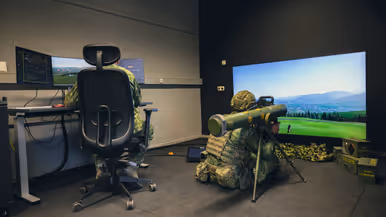 Een viertal afbeeldingen. Naast het cilindervormige, donkergroene antitankwapen Spike Long Range gericht op een beamer, hetzelfde middel alleen nu met daarachter een militair. Samen met een instructeur, die achter een beeldscherm zit, bevinden de twee zich in een kleinere ruimte. Vervolgens diezelfde oefenende militair schuin vanaf onder en gezien vanaf voren. Daarbij is zijn geconcentreerde blik zichtbaar.