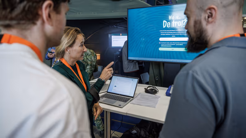 Drie personen voeren een gesprek voor een laptopscherm en een groot beeldscherm.