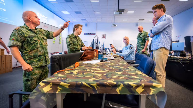 Een tafel met daaromheen zowel militairen als burgers. Kolonel Patrick wijst daarbij.