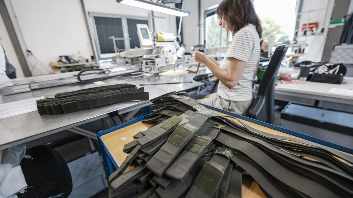 Stapel bretels in camouflagepatroon in een atelier. Daarvoor een vrouw die aan een bureau werkt met deze bretels.