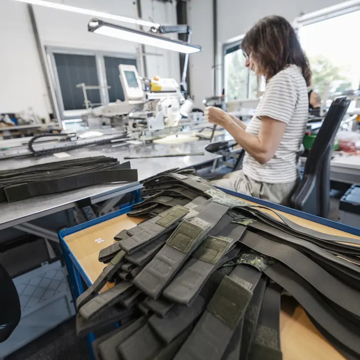 Stapel bretels in camouflagepatroon in een atelier. Daarvoor een vrouw die aan een bureau werkt met deze bretels.