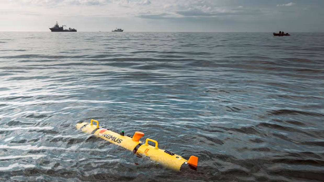 Marinecollega’s met een bouwvakkershelm op zijn bezig met een zeedrone die in de takels hangt. Op de andere foto ligt de gele zeedrone in het water, op de achtergrond zijn marineschepen te zien. 