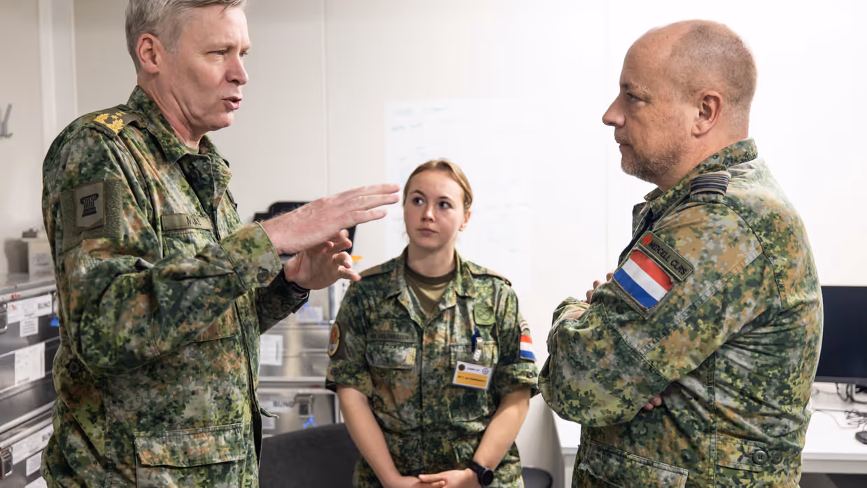 Links: Drie militairen in uniform staan in gesprek. Een man wijst uitleggend naar voren, een vrouw luistert aandachtig, en een andere man staat met gekruiste armen. Rechts: Een vrouw in militair uniform met naam praat serieus tijdens een bespreking. Op de achtergrond staan collega's in uniform bij een whiteboard. 