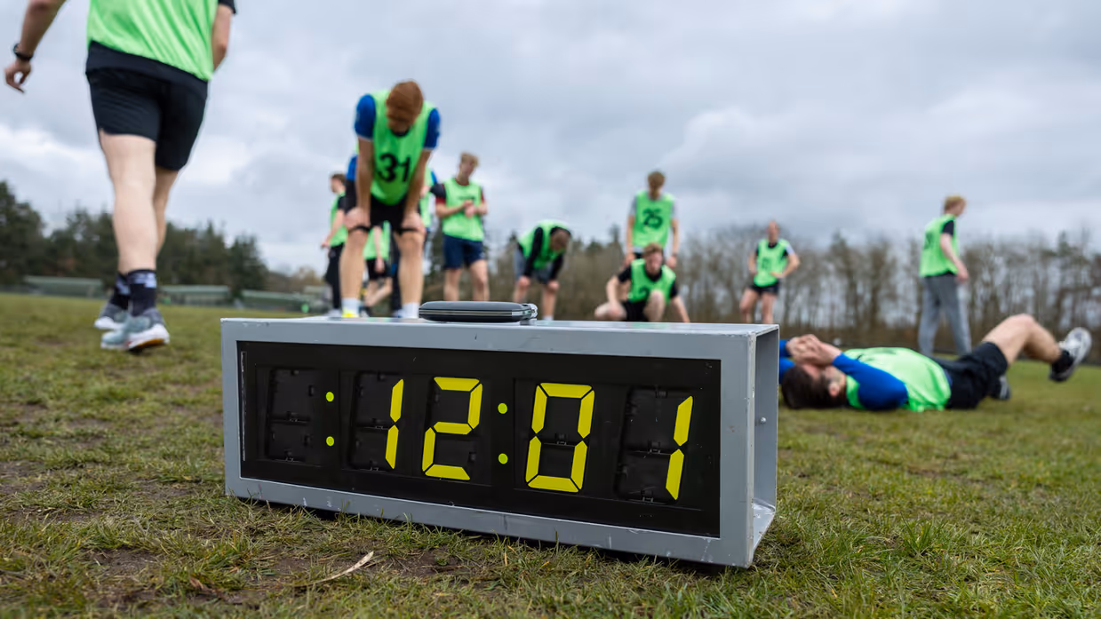 Een groep hardlopers in gele hesjes met nummers rent op een atletiekbaan. De voorste loper lijkt gefocust, met bomen en een hek op de achtergrond.