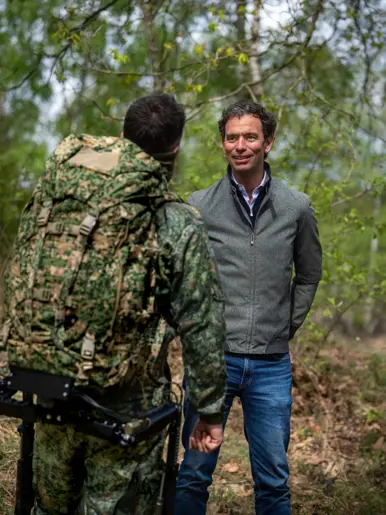 Een man in grijs jack praat met een soldaat in camouflage en rugzak in een groen bos. 