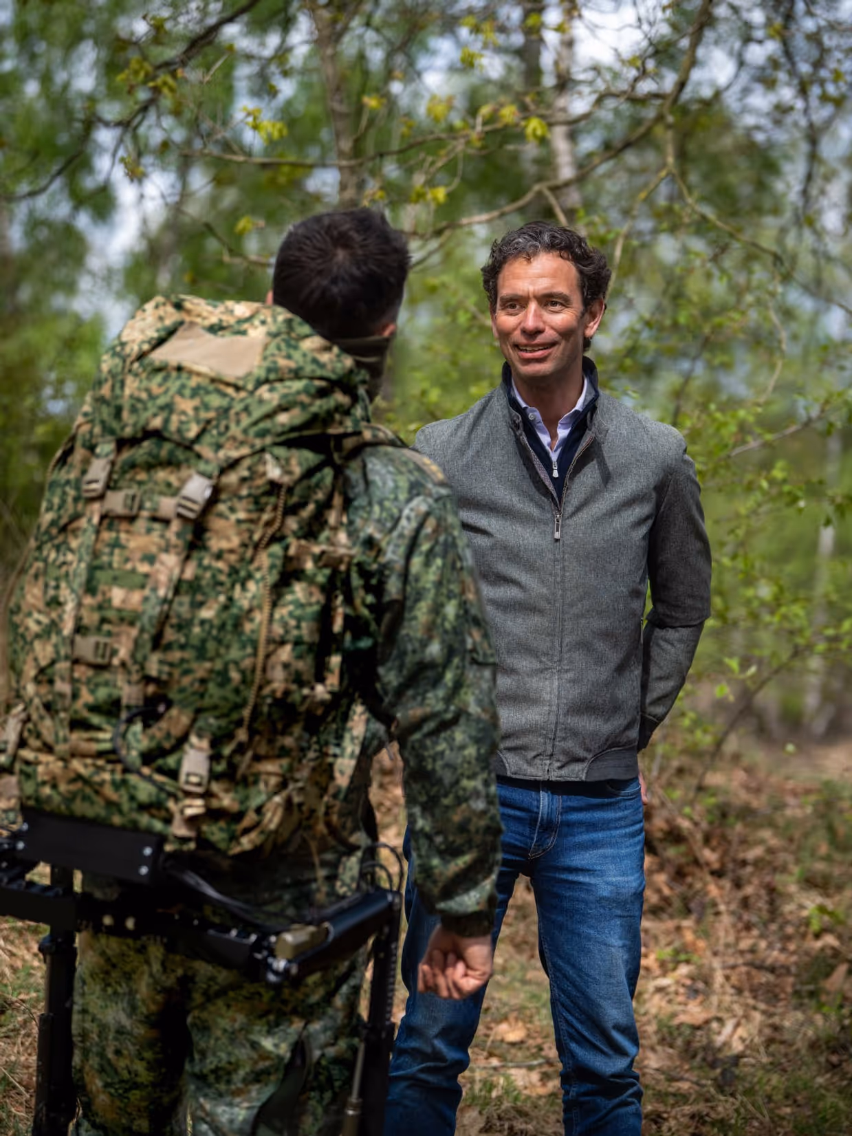 Een man in grijs jack praat met een soldaat in camouflage en rugzak in een groen bos. 