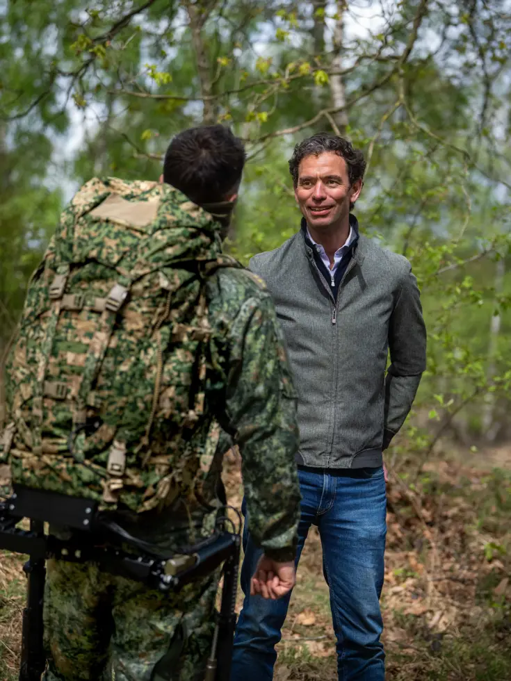 Links: Twee mannen in uniform: één met camouflage, rugzak en exoskelet, de andere in zwart met kogelwerend vest. Rechts: Een man in grijs jack praat met een soldaat in camouflage en rugzak in een groen bos. 