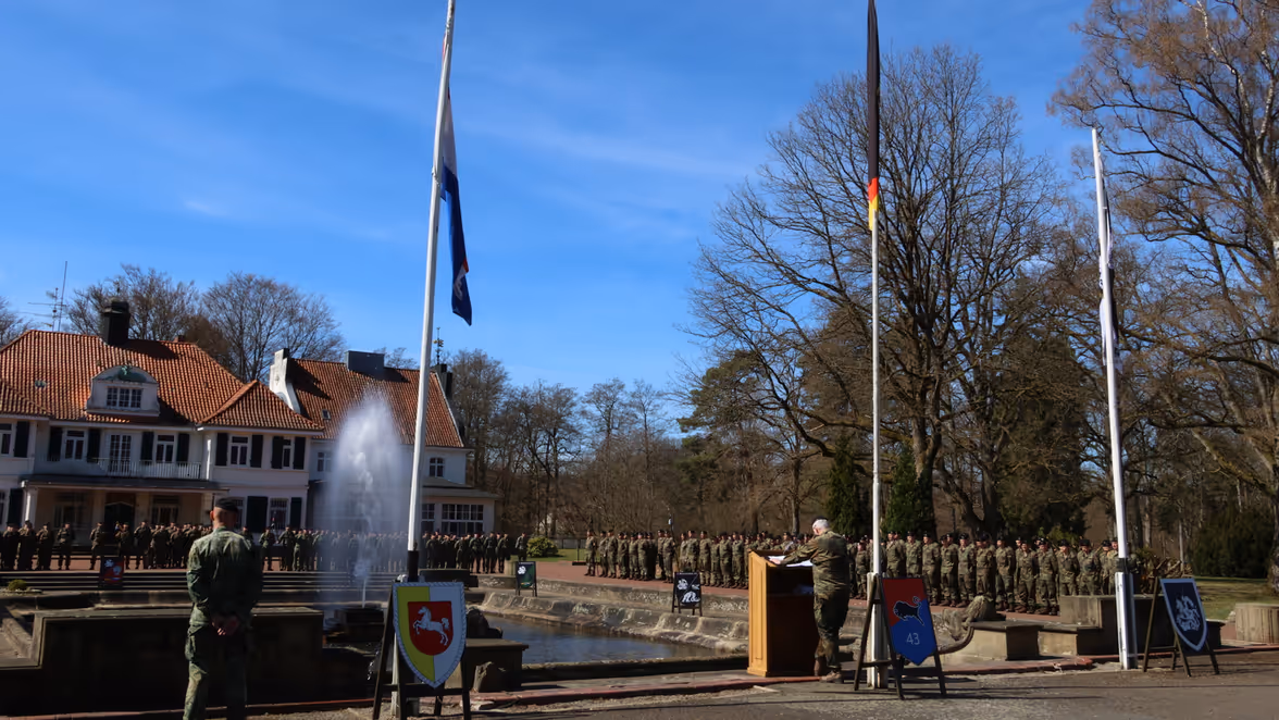 Een militair staat achter een spreekgestoelte met de Nederlands en Duitse vlag in top, met veel militairen in gelid op de achtergrond.