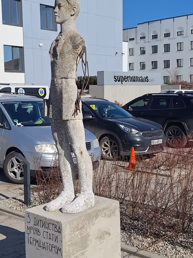 Een groot, liggend stenen beeld van een persoon, half ingebed in een betonnen blok, naast een modern gebouw met geparkeerde auto’s en een heldere lucht.