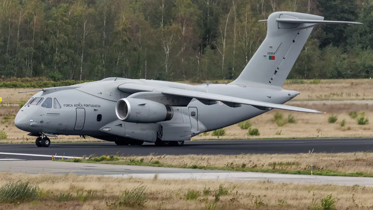  Een grijs militair transportvliegtuig van de Portugese luchtmacht rollend op een startbaan met gras, bomen en een bewolkte lucht. 