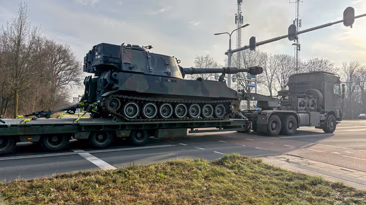 Een groene pantserhouwitser staat op een lage oplegger, getrokken door een zware vrachtwagen op een weg met verkeerslichten. 