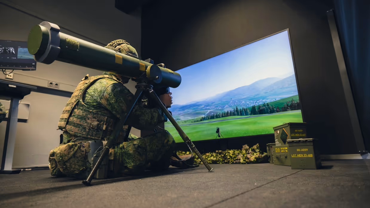 Een viertal afbeeldingen. Naast het cilindervormige, donkergroene antitankwapen Spike Long Range gericht op een beamer, hetzelfde middel alleen nu met daarachter een militair. Samen met een instructeur, die achter een beeldscherm zit, bevinden de twee zich in een kleinere ruimte. Vervolgens diezelfde oefenende militair schuin vanaf onder en gezien vanaf voren. Daarbij is zijn geconcentreerde blik zichtbaar.