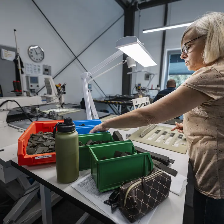  Een vrouw achter een tafel in een atelier pakt onderdelen uit het bakje voor het in elkaar zetten van bretels.