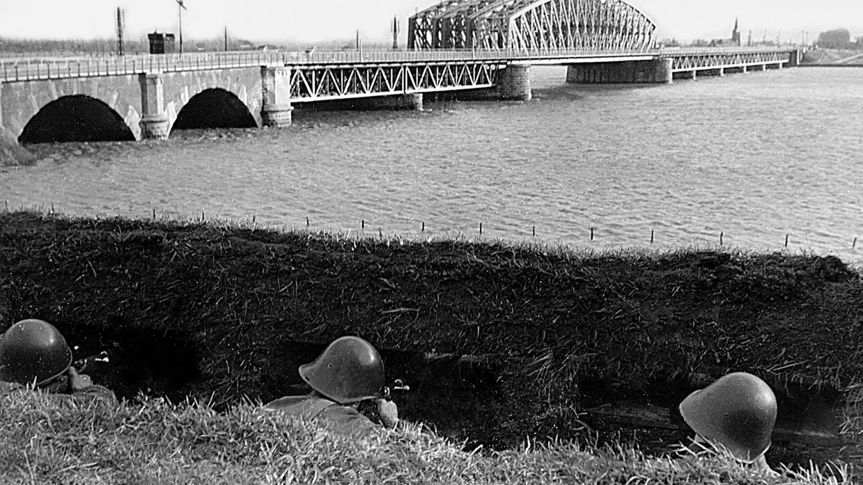 Zwart-wit foto van 3 militairen tijdens Meidagen 1940. Ze liggen in dekking bij brug over water.