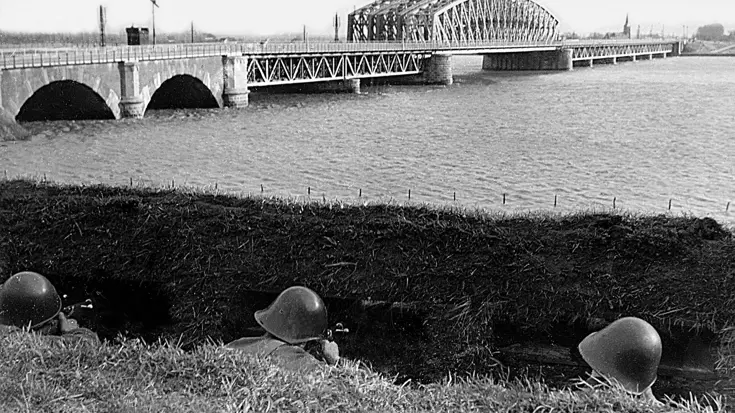 Zwart-wit foto van 3 militairen tijdens Meidagen 1940. Ze liggen in dekking bij brug over water.