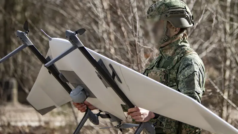 Een militair loopt door het terrein met een grote drone in zijn handen. 