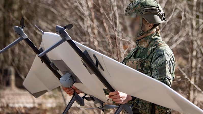 Een militair loopt door het terrein met een grote drone in zijn handen. 