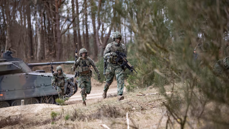 Gewapende militairen rennen naar de bosrand. Op de achtergrond een CV90.   