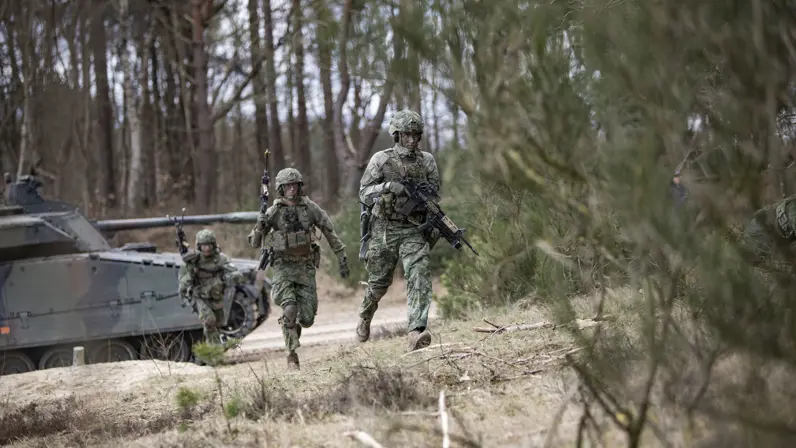 Gewapende militairen rennen naar de bosrand. Op de achtergrond een CV90.   
