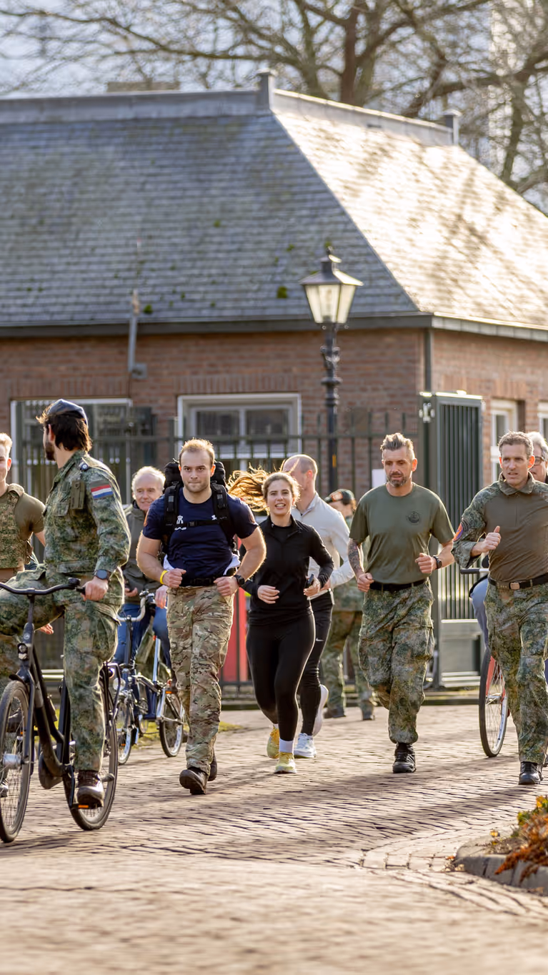 Een groep mensen in beweging op een zonnig pad: sommigen lopen, anderen fietsen. Ze dragen sportieve kleding en militaire uniformen, met bomen en een gebouw op de achtergrond.