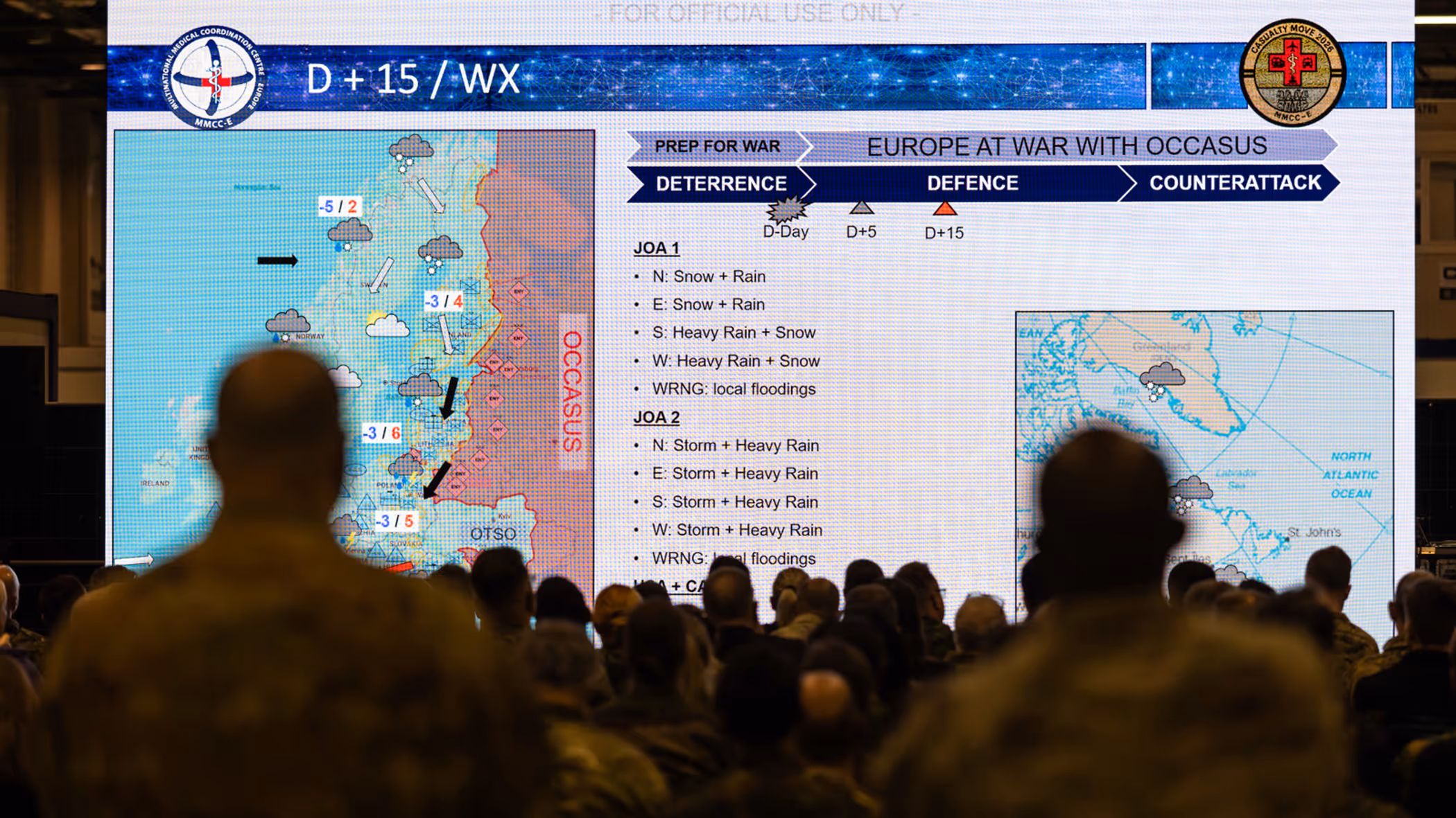 Een militaire presentatie met een groot scherm. Op het scherm staat een weerkaart van Europa met tekst over "Europa in oorlog" en weeromstandigheden. Een groep mensen kijkt toe.