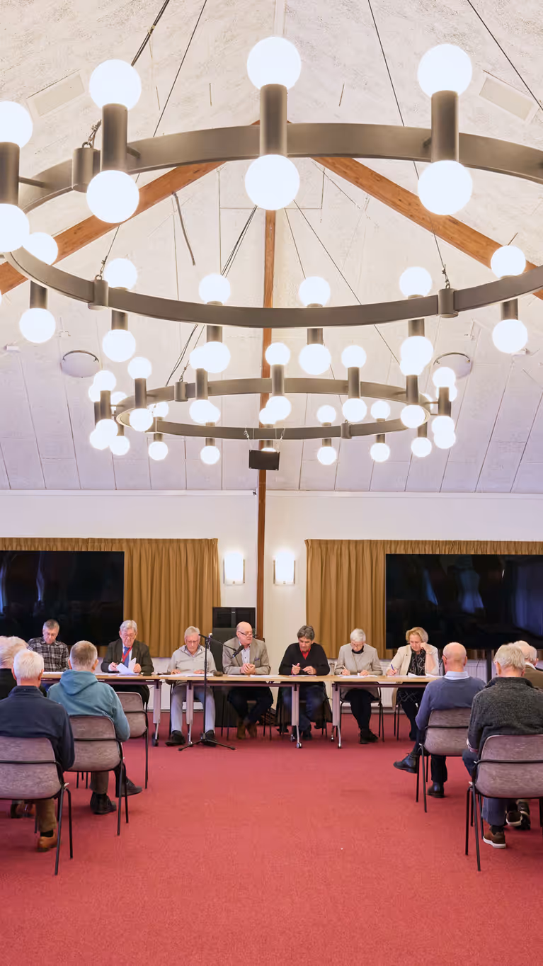 In een oude zaal zitten oudere mensen en achter de tafel een man met een hamer en serieus kijkende mensen.