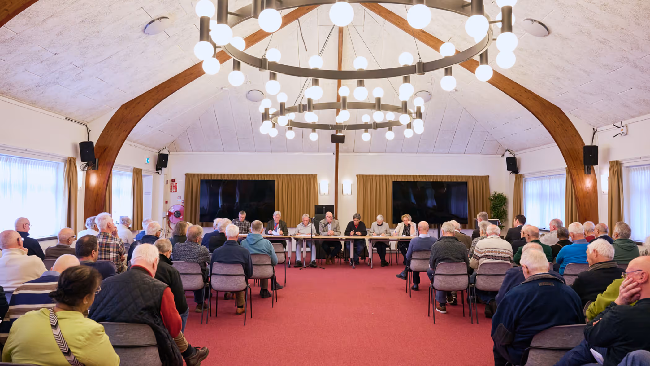 In een oude zaal zitten oudere mensen en achter de tafel een man met een hamer en serieus kijkende mensen.