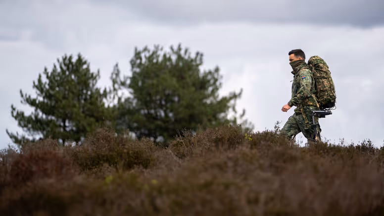 Een soldaat in camouflagekleding met een grote rugzak loopt door een heideveld met dennenbomen op de achtergrond. 
