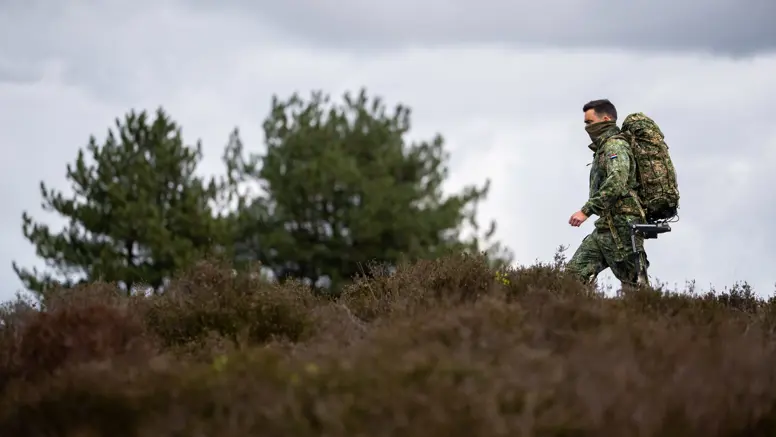Een soldaat in camouflagekleding met een grote rugzak loopt door een heideveld met dennenbomen op de achtergrond. 