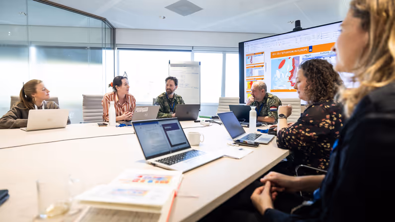 Een kamer met tafels waaraan burgers en militairen zitten met laptops voor zich. 