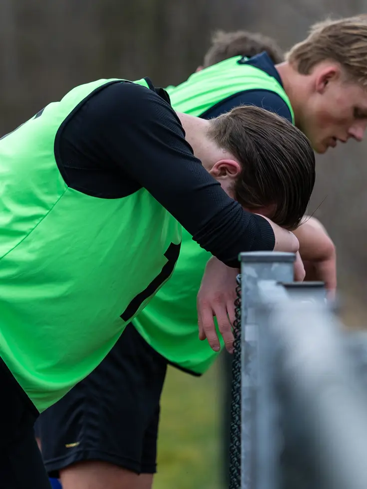 Een groep hardlopers in gele hesjes met nummers rent op een atletiekbaan. De voorste loper lijkt gefocust, met bomen en een hek op de achtergrond.