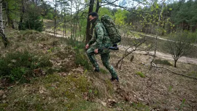Een militair in camouflage met rugzak en exoskelet loopt door een bos met bomen en struiken op een heuvel.