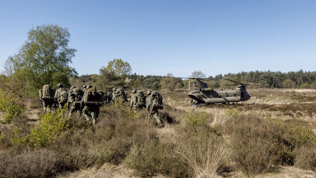 Een groep soldaten in camouflage loopt door een heideveld met lage struiken, een helikopter staat op de achtergrond. 