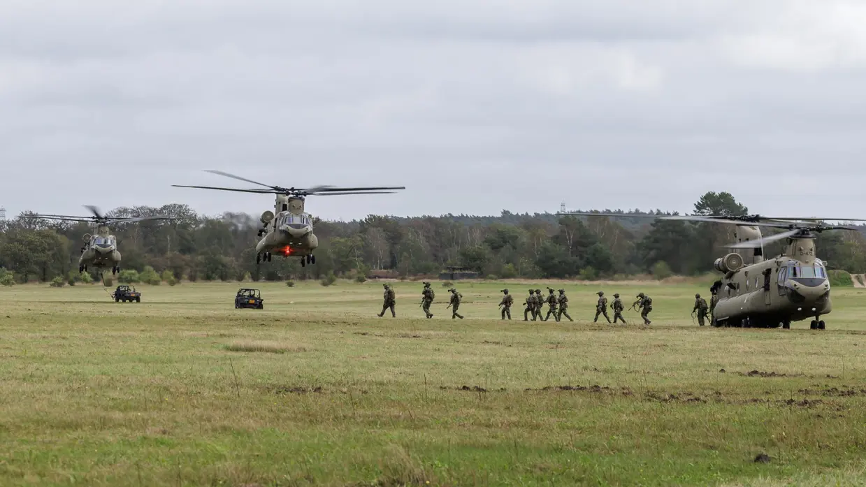 Drie grote militaire helikopters landen in een groen veld, soldaten rennen uit de voorste, voertuigen en bomen op de achtergrond.