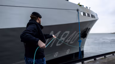 Een militair van de marine haalt met een touw een van de trossen binnen. Daarnaast enkele matrozen en onderofficieren bij de reling op het dek van het schip.