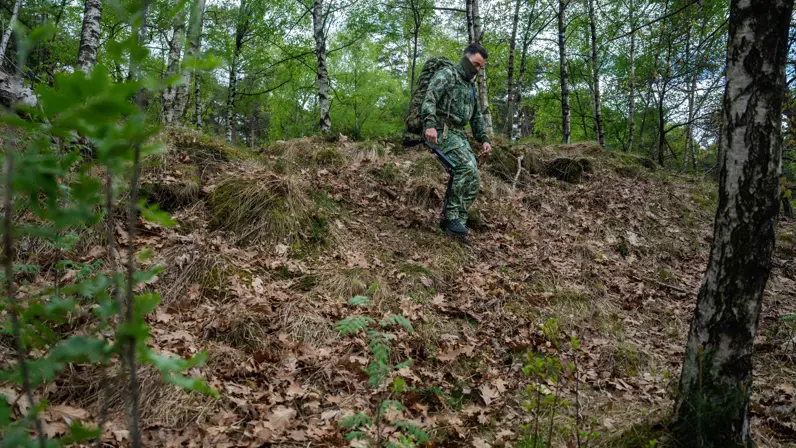 Links: Een soldaat in camouflage met rugzak en exoskelet loopt door een bos met bomen en struiken op een heuvel.    Rechts: een soldaat in camouflage met rugzak en stokken klimt een met bladeren bedekte heuvel op in een groen bos. 