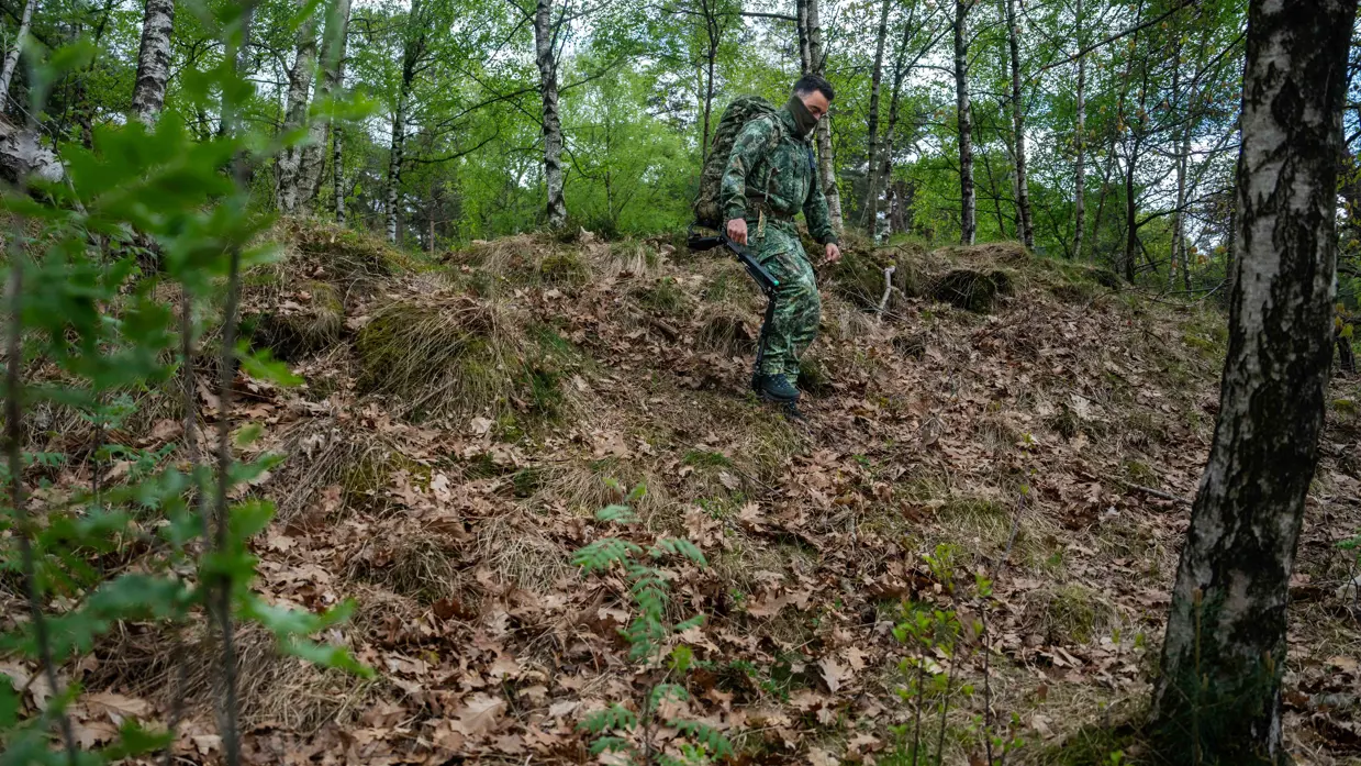 Een militair in camouflage met rugzak en stokken klimt een met bladeren bedekte heuvel op in een groen bos. 
