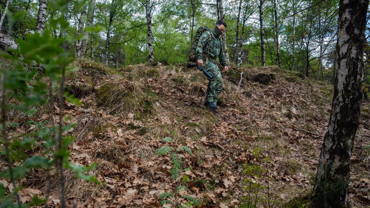 Links: Een soldaat in camouflage met rugzak en exoskelet loopt door een bos met bomen en struiken op een heuvel.    Rechts: een soldaat in camouflage met rugzak en stokken klimt een met bladeren bedekte heuvel op in een groen bos. 